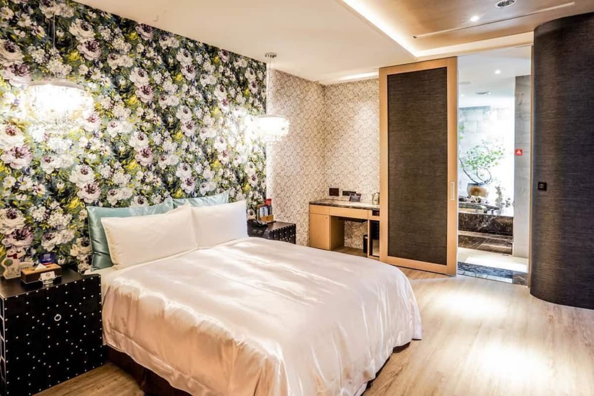 A bedroom with a floral wallpaper, a bed with white bedding, a desk with a chair, and a view of an outdoor area.