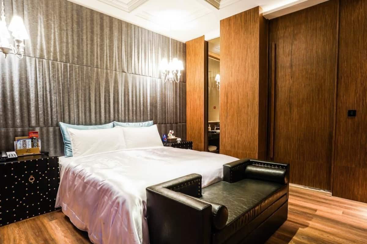 A modern hotel room with a large bed, a leather sofa, wooden paneling, and a chandelier.
