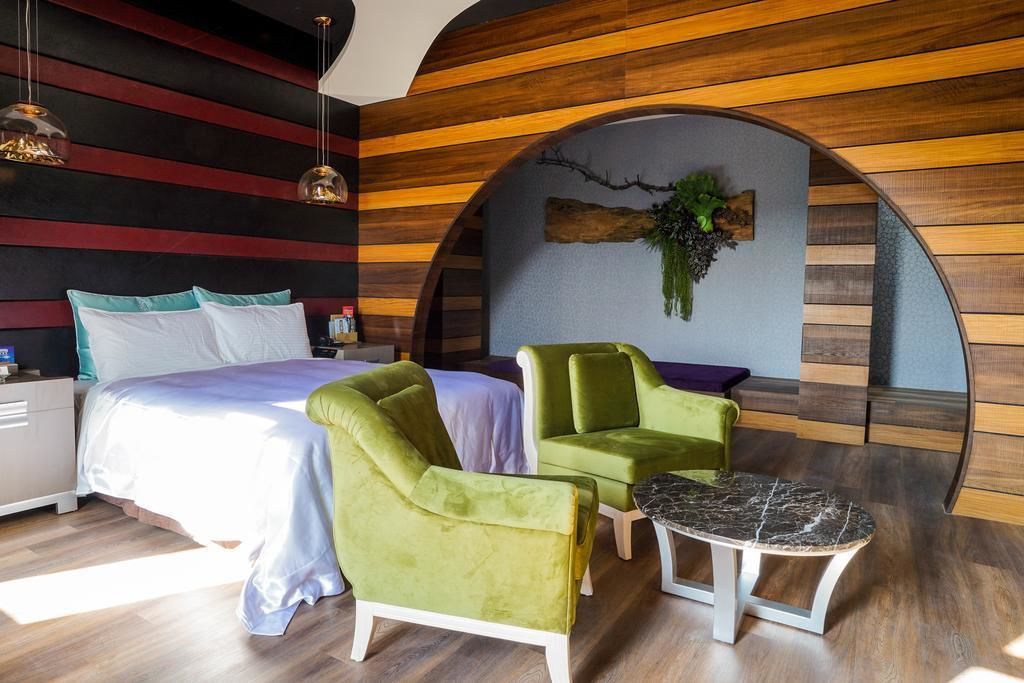 A modern bedroom with a bed, two green armchairs, a small table, and a unique curved wooden wall design.