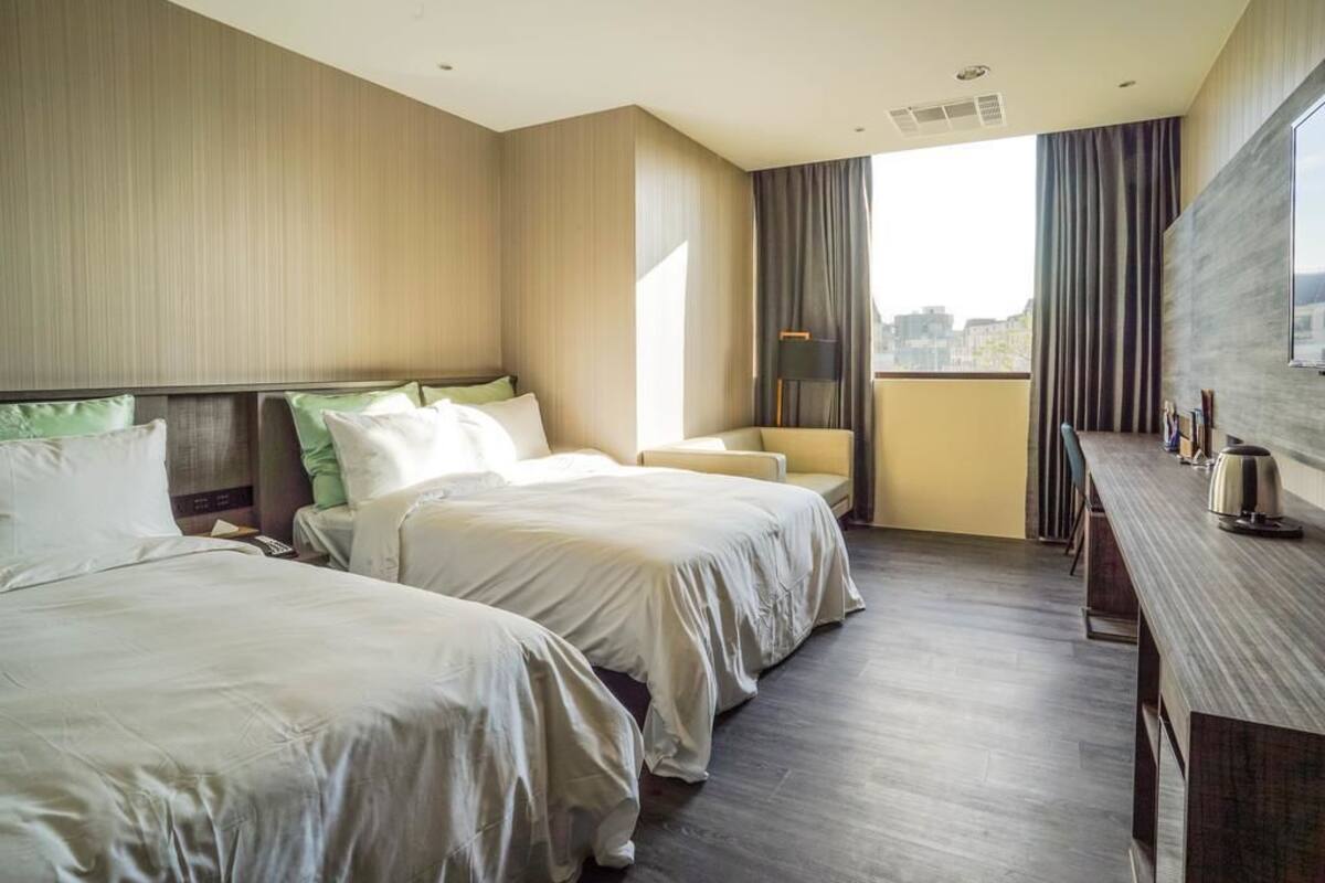 A hotel room with two beds, a desk, a chair, and a large window with curtains.