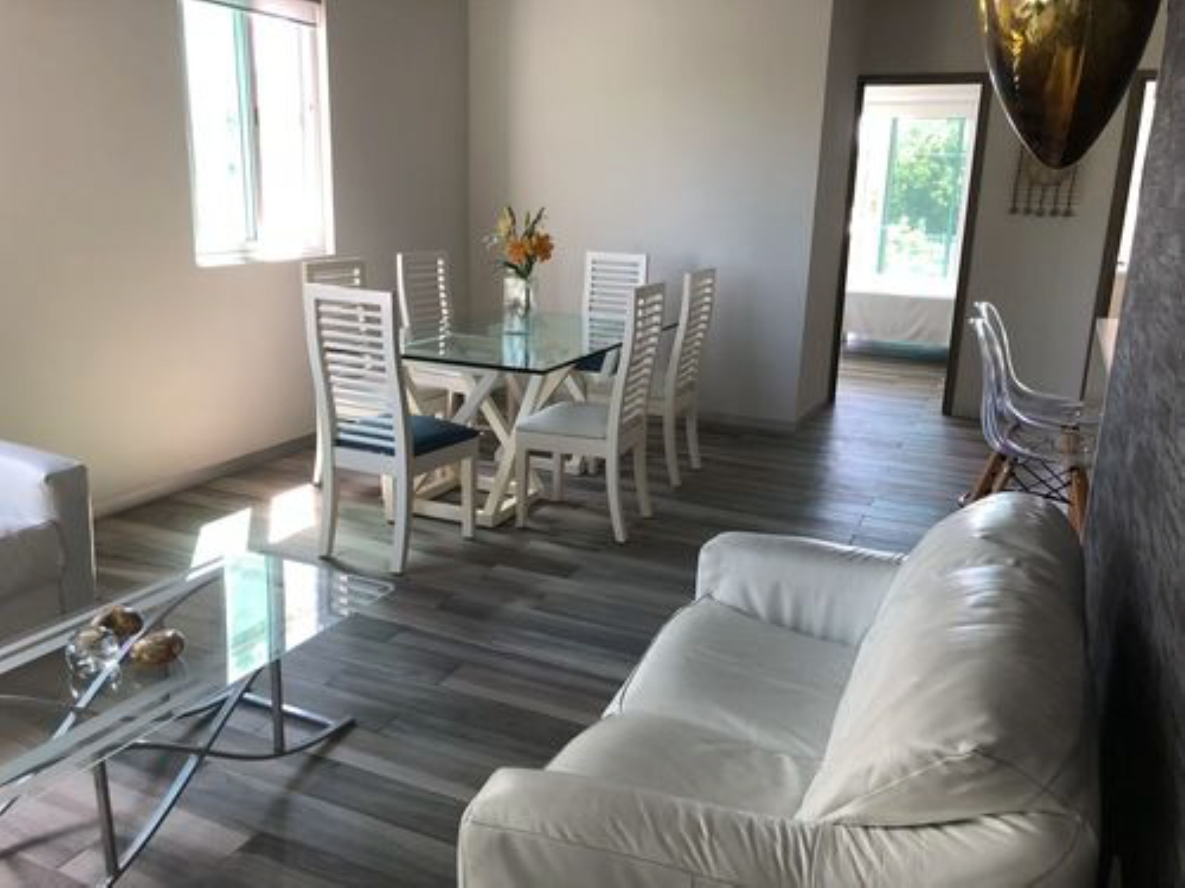 A modern living room with a white sofa, a glass coffee table, and a dining area with white chairs and a glass table.