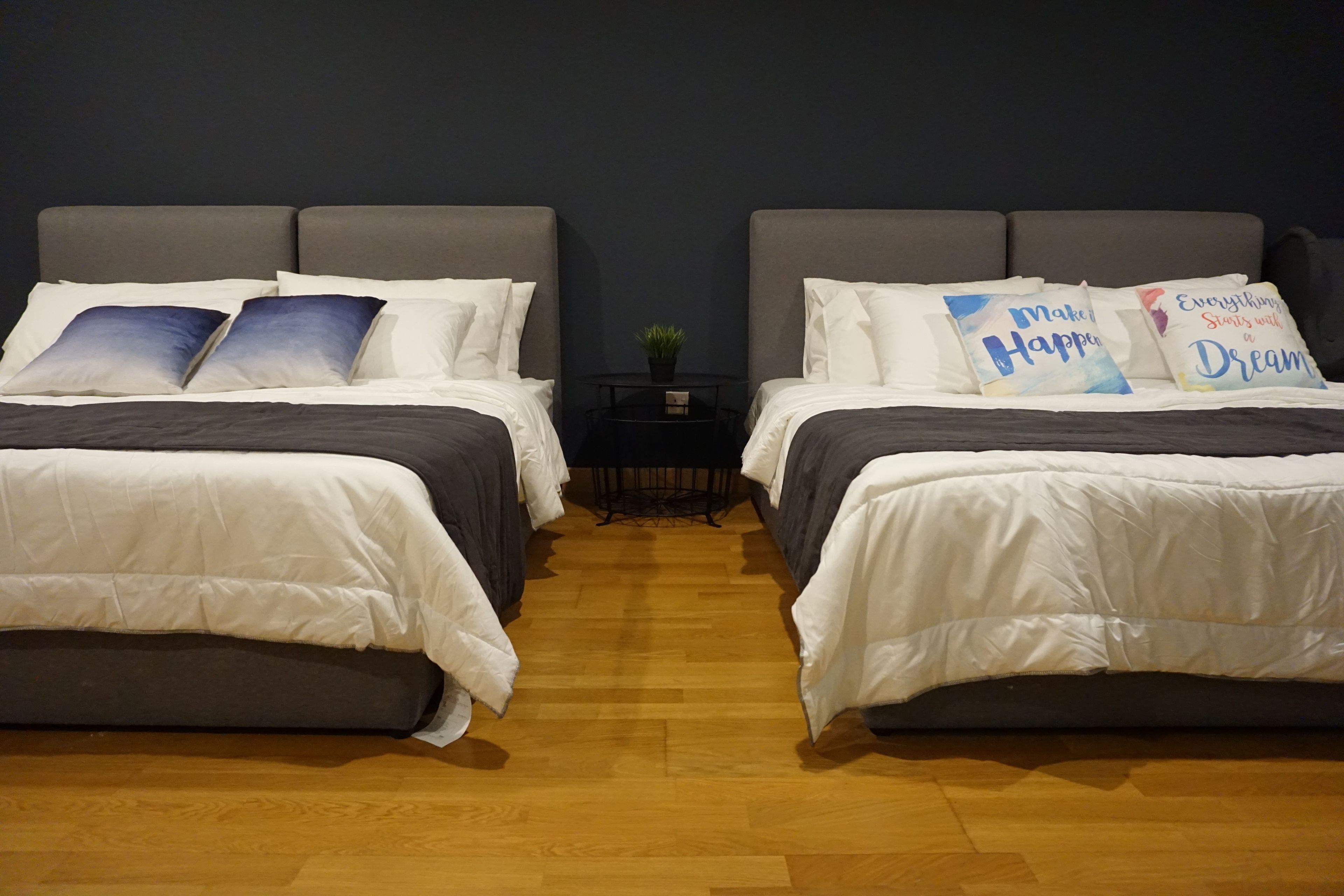 A room with two beds, each with white bedding and grey headboards. There are decorative pillows with motivational quotes. A small table with a plant is between the beds.