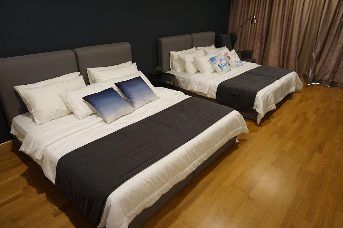 A hotel room with two beds, each with white bedding and a dark gray headboard. There is a small side table with a lamp and a decorative pillow. The room has wooden flooring and a dark blue wall.