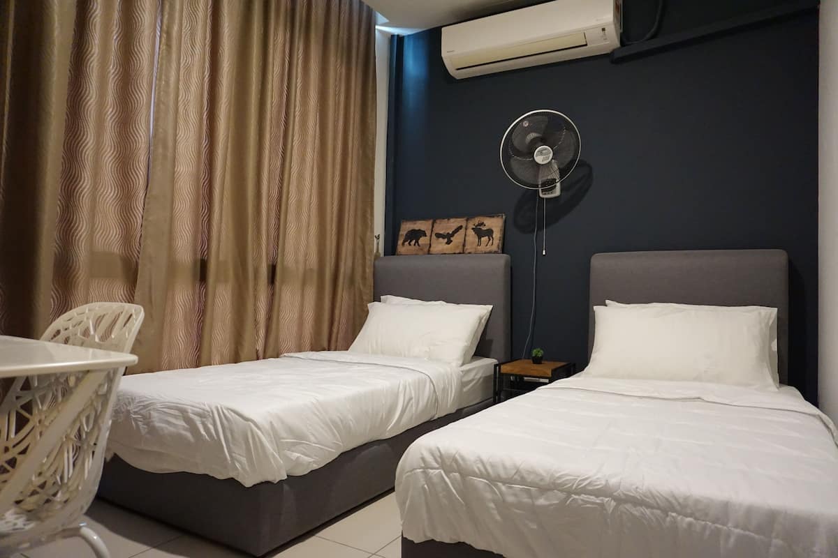 A hotel room with two beds, a table, a chair, an air conditioner, a wall-mounted fan, and a decorative wooden panel.