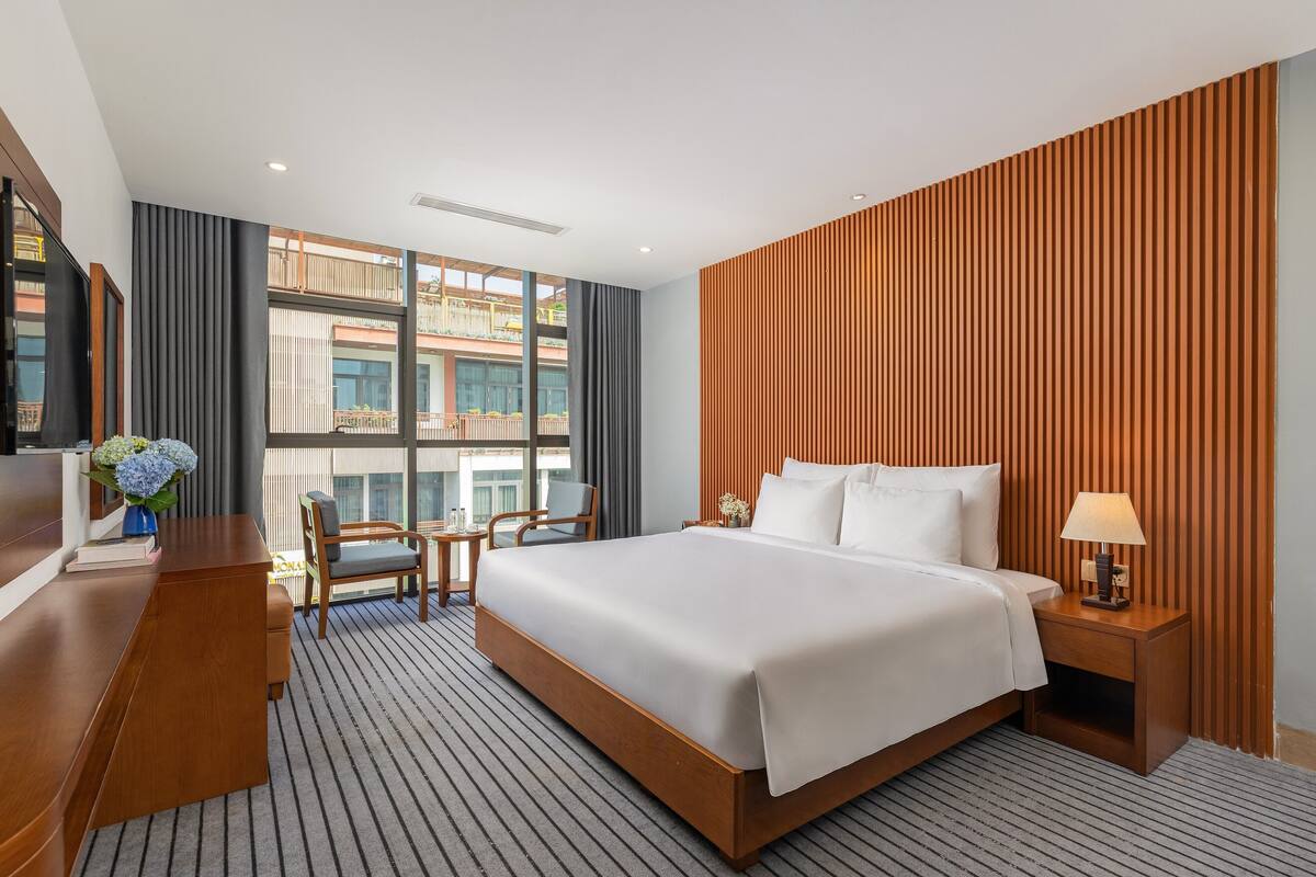 Habitación de hotel con una cama grande, un escritorio, una silla, una mesita y una ventana con vistas a edificios.