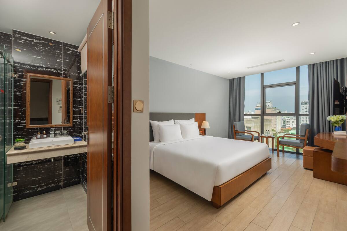 Habitación de hotel con una cama grande, un baño con tocador de mármol y vistas a la ciudad a través de amplias ventanas.