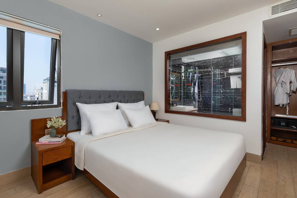 Una cama bien hecha con ropa blanca, un cabecero tapizado en gris y una mesita de noche con un jarrón de flores y libros.