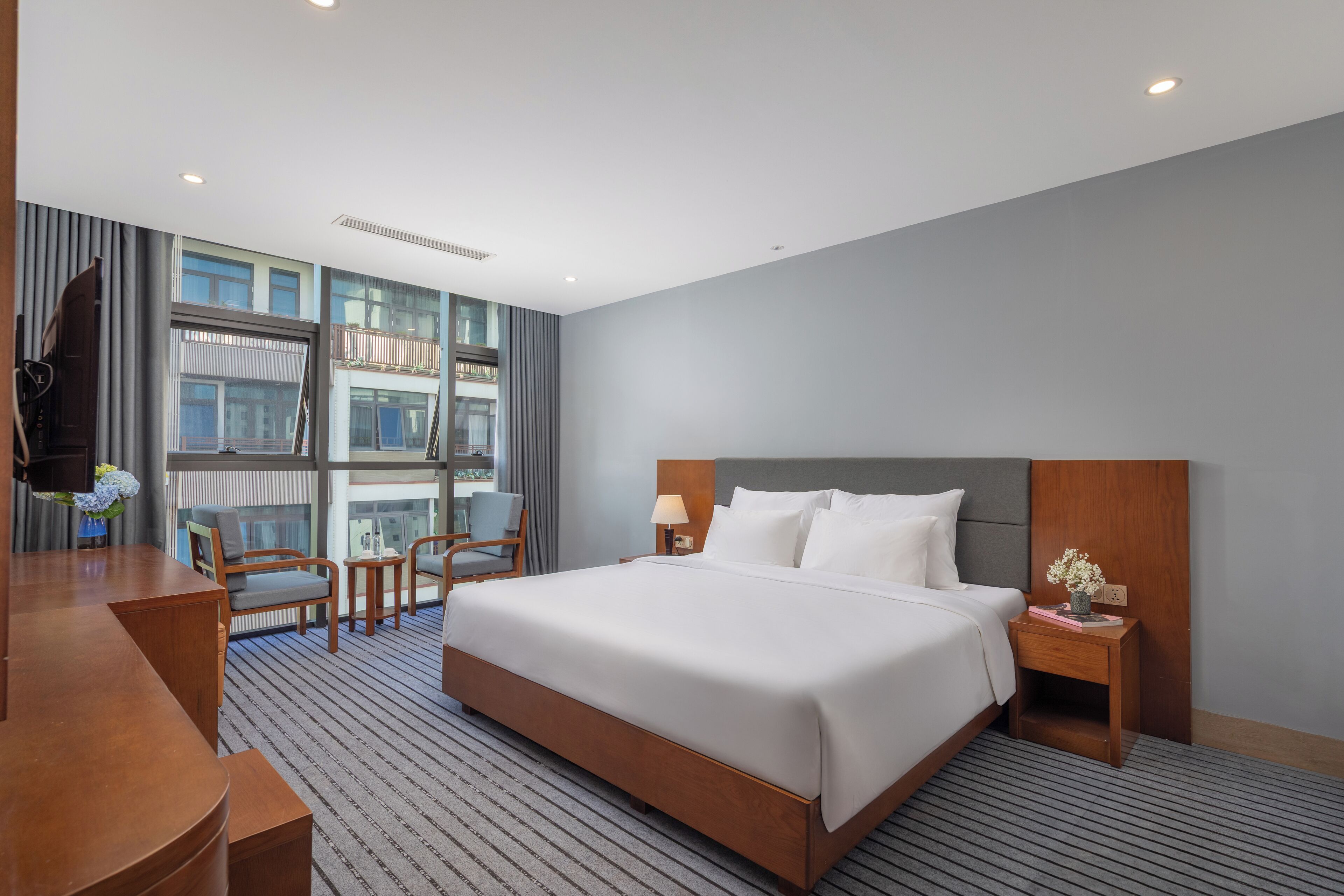 A hotel room with a large bed, a desk, and a chair. There is a window with a view of buildings.