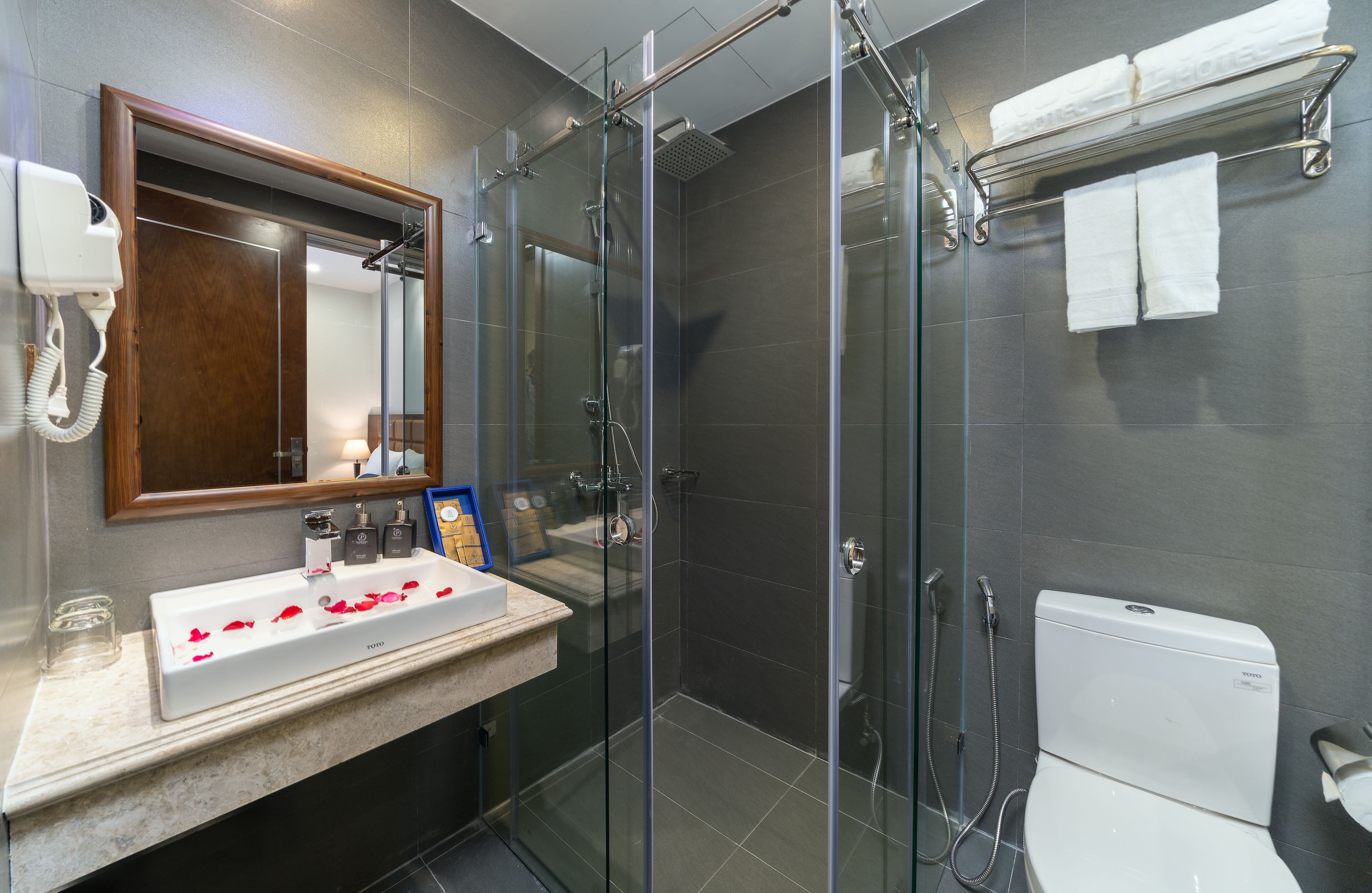 A modern bathroom with a glass-enclosed shower, a white sink with rose petals, a hairdryer, and a toilet.