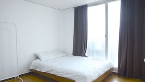 Quarto individual com uma cama, piso de madeira, paredes brancas e uma janela grande com cortinas cinzentas.