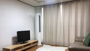 Uma sala de estar moderna com uma televisão de tela plana sobre um suporte de madeira, um tapete felpudo branco, um sofá bege e uma janela grande com cortinas leves.