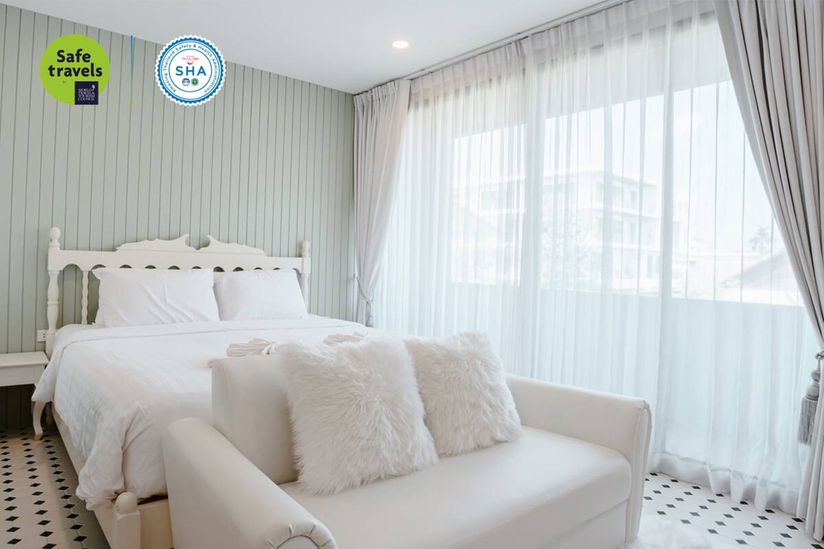 A bedroom with a bed, a white sofa, and a window with sheer curtains.