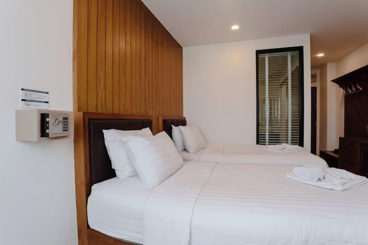 A hotel room with two beds, a wooden panel wall, a window with blinds, and a control panel on the wall.