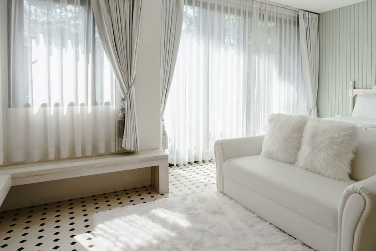 A bedroom with a bed, a white sofa with fluffy pillows, a bench, and sheer curtains.