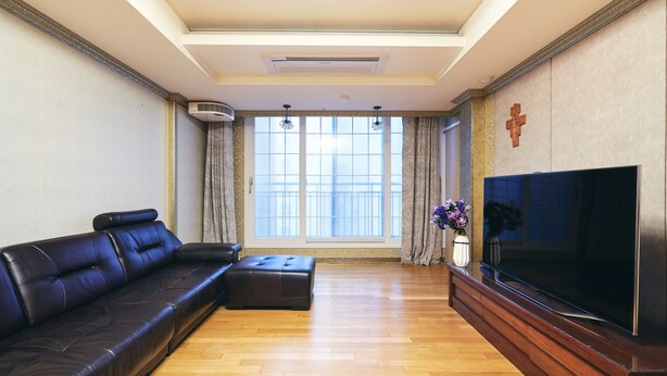Ein Wohnzimmer mit einem schwarzen Ledersofa, einem Couchtisch aus Holz, einem Flachbildfernseher und einem großen Fenster mit Vorhängen.