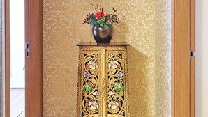 Ein Eckzimmer mit ornamentalem Wandpapier, Holzboden und einem dekorativen Schrank mit Blumen in einer Vase.