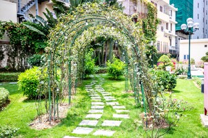 Un sendero de jardín con pavimento de piedra, rodeado de follaje y un arco cubierto de plantas trepadoras.