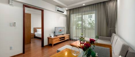 Una habitación de hotel moderna con una mesa de comedor de madera, un sofá, un televisor y un ventanal con cortinas.