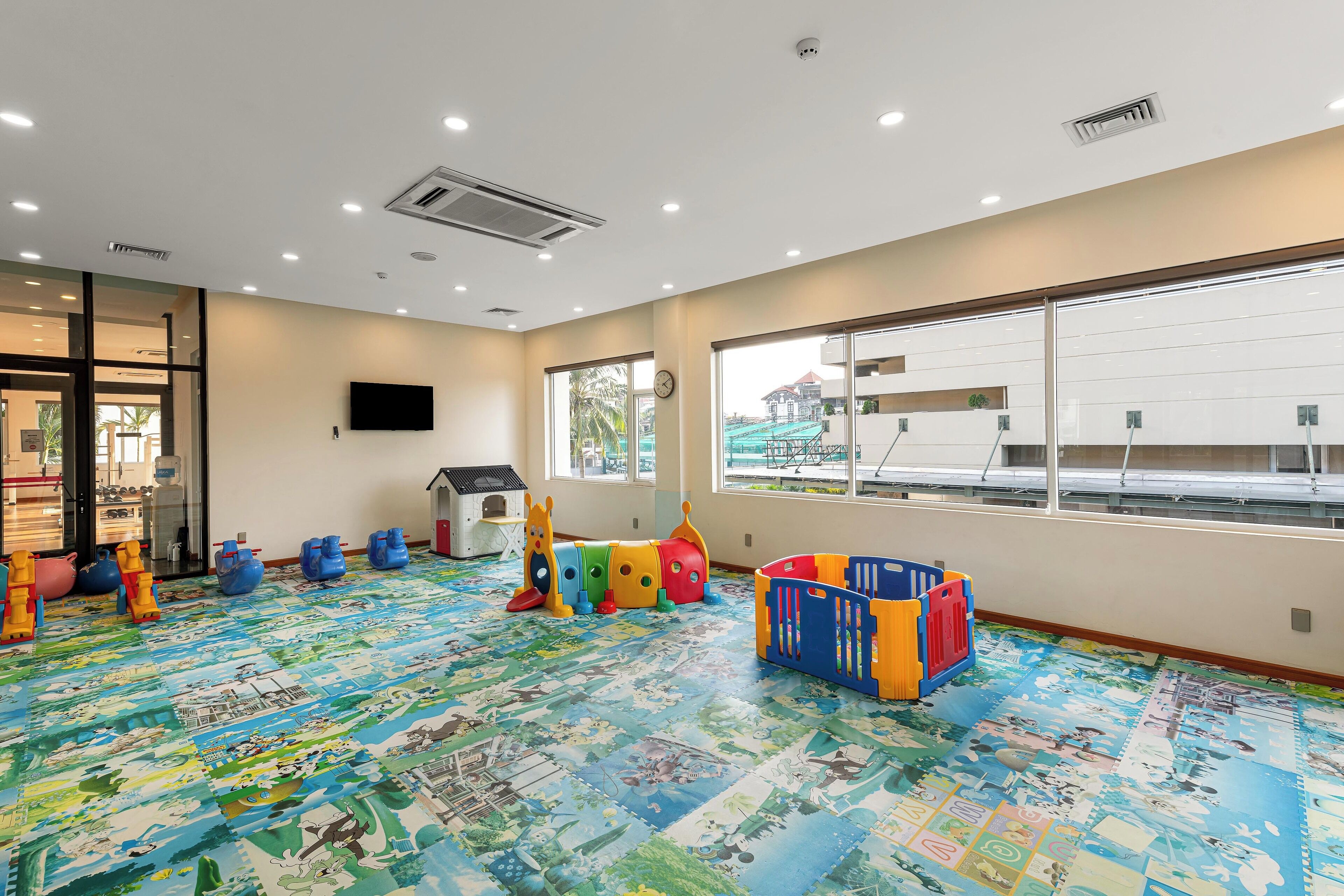 A spacious room with a colorful play mat, a large window, and a flat-screen TV on the wall.