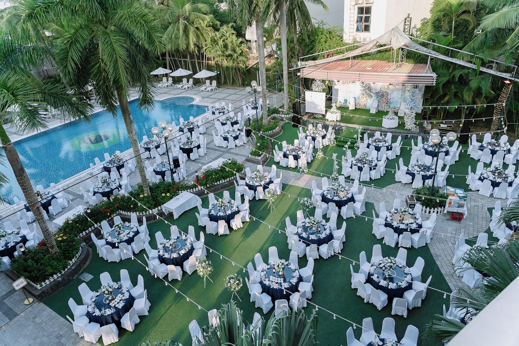 Een evenementenlocatie buiten met ronde tafels en stoelen, een zwembad en palmbomen.