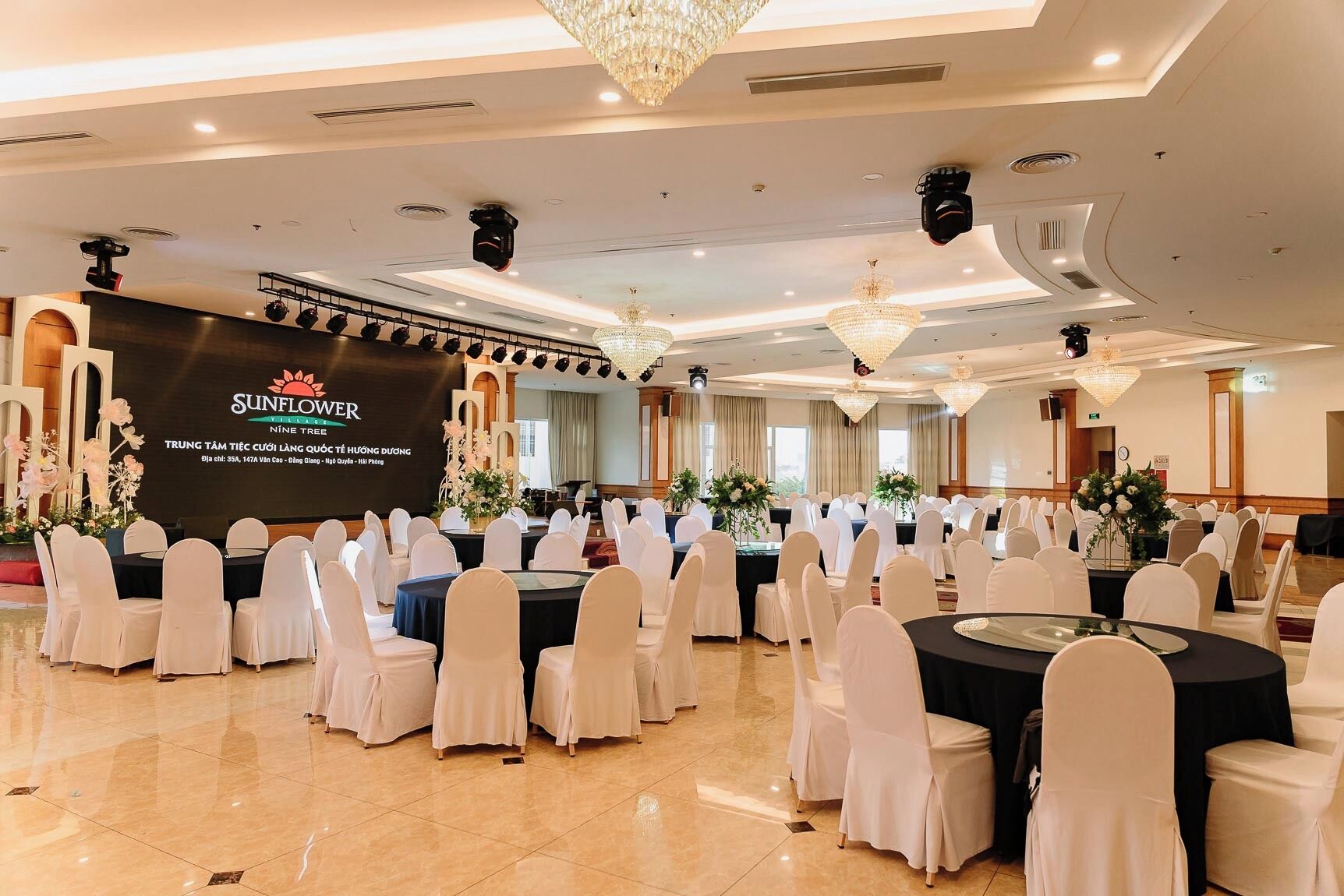 A conference room with round tables and white chairs, a stage with a backdrop, chandeliers, and a polished floor.