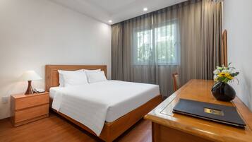 A hotel room with a wooden bed, a bedside table with a lamp, a desk with a laptop, and a window with curtains.