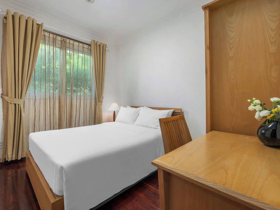 Una camera d'albergo con un letto, una testiera in legno, un comodino con una lampada, un tavolo in legno e un vaso con fiori.