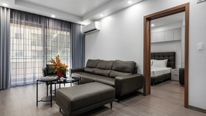 A modern living room with a sectional sofa, a small coffee table, and a large ottoman. There is a bed with a headboard in the adjacent room. - Sunflower International Village (Hai Phong)