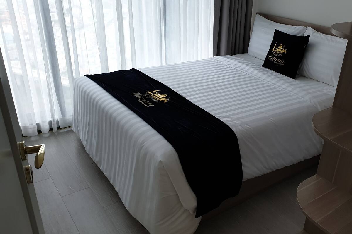 Une chambre d’hôtel avec un grand lit, du linge de lit blanc, un plaid noir et un coussin noir orné d’un motif.