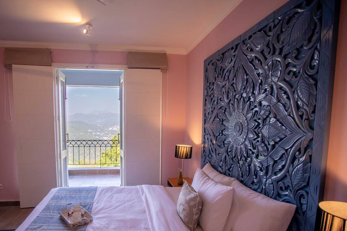 Une chambre d’hôtel avec un grand lit, une tête de lit décorative, un balcon avec vue et un plateau avec des tasses et une théière posé sur le lit.