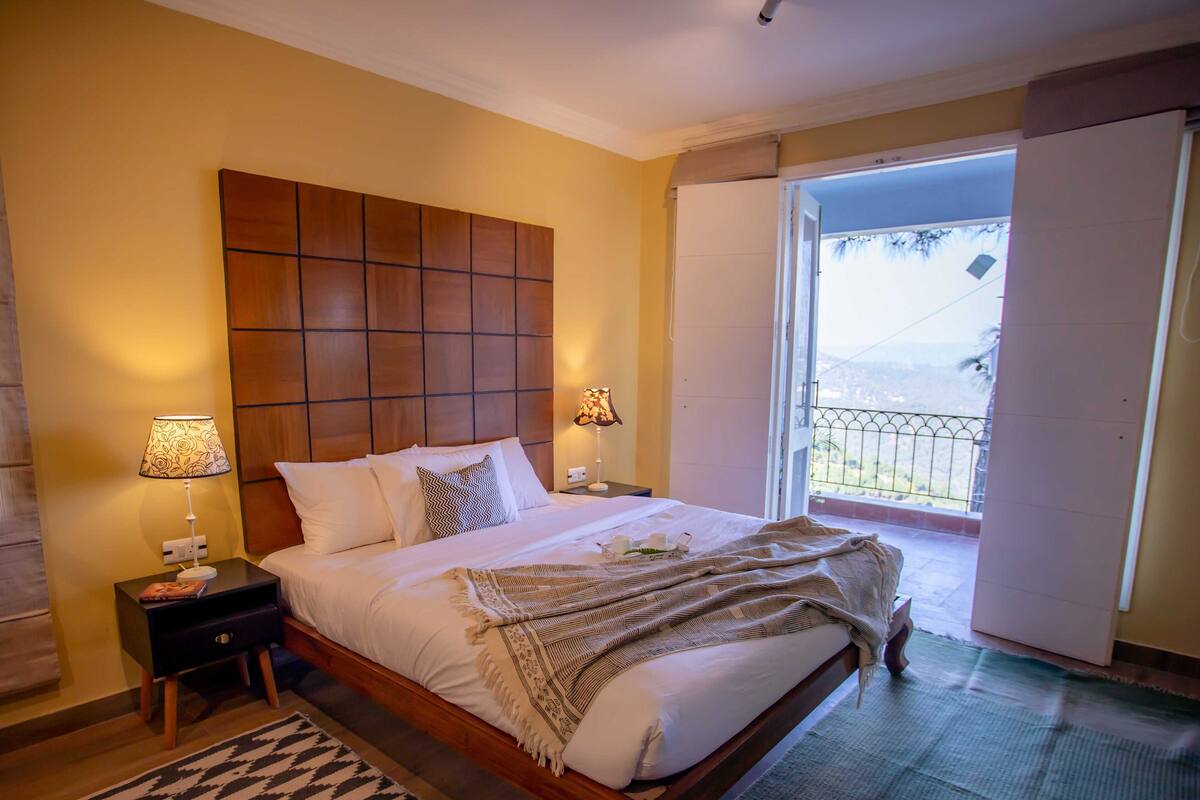 Un lit bien fait, avec une tête de lit en bois, deux lampes de chevet, un tapis à motifs et une vue sur un balcon doté d’une balustrade.