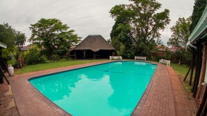 Una piscina rectangular con borde de ladrillo, rodeada por un sendero de ladrillo y una estructura con techo de paja al fondo.