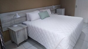 Una cama bien hecha con colcha blanca acolchada, cabecera de madera y mesita de noche con lámpara.