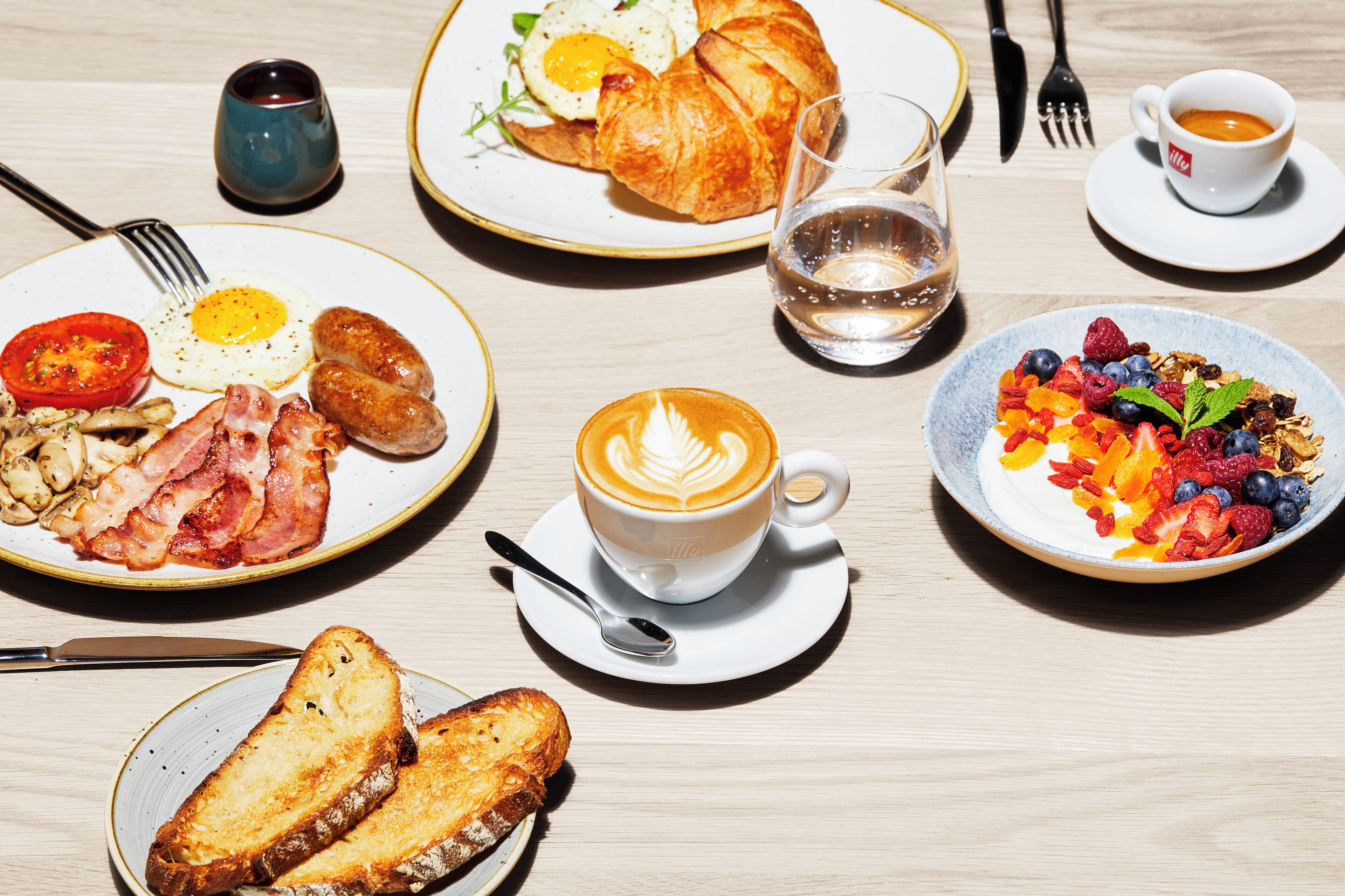 A breakfast setting with plates of eggs, bacon, sausages, toast, and a coffee drink.