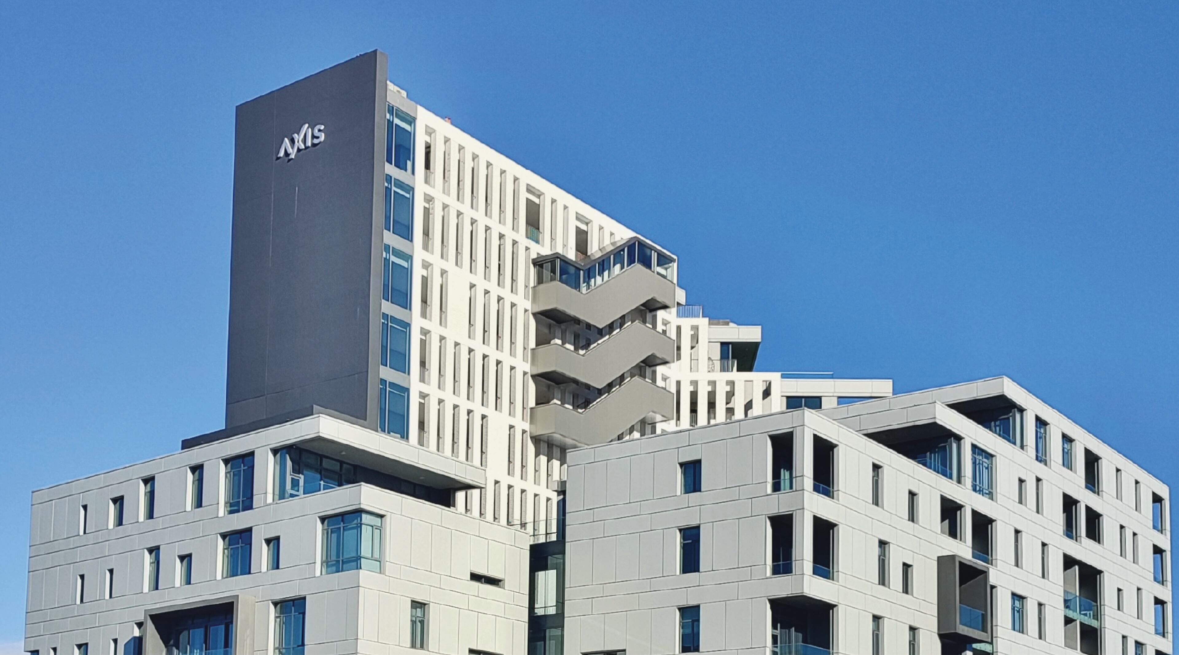 Ein modernes Gebäude mit einem gut sichtbaren 'AXIS'-Schild, mehreren Balkonen und wolkenlosem, blauem Himmel.