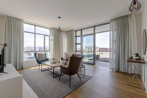 Uma sala de estar moderna com um sofá, poltronas, uma mesa de centro e uma TV. Janelas amplas oferecem vista para a cidade.