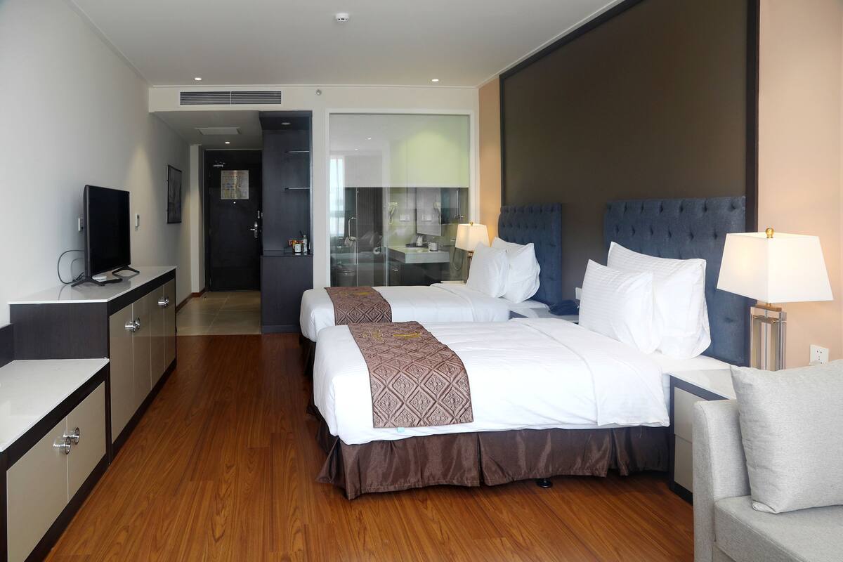 Ein Hotelzimmer mit zwei Betten, einem Fernseher, einem Schreibtisch und einem durch eine Glastür einsehbaren Badezimmer.