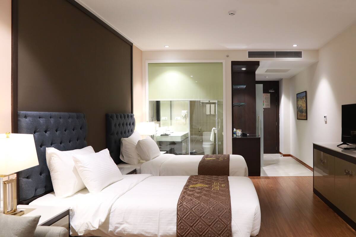 Ein Hotelzimmer mit zwei Betten, einem großen Spiegel, einem Fernseher und einem durch eine Glastür einsehbarer Badezimmer.