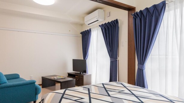Une chambre d’hôtel avec un lit, un téléviseur posé sur un meuble, un fauteuil bleu et une petite table avec un magazine.