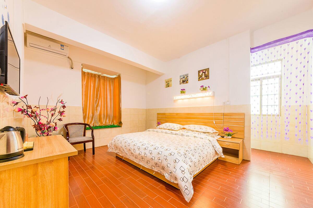 Quarto de hotel com uma cama, uma mesa com chaleira, uma cadeira, uma janela com cortinas e obras de arte na parede.