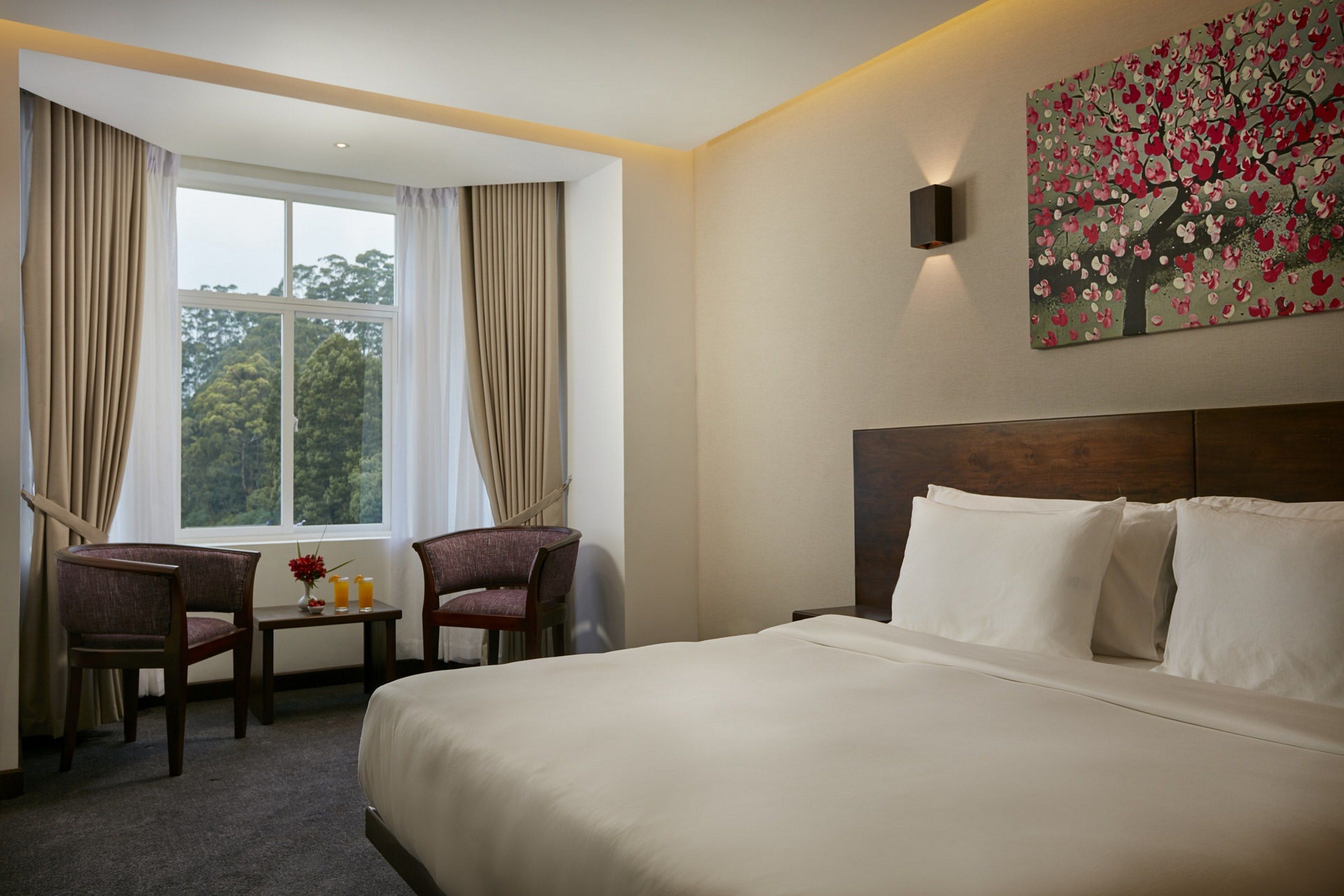 A hotel room with a large bed, two chairs, a small table with a vase and two glasses, and a window with curtains.