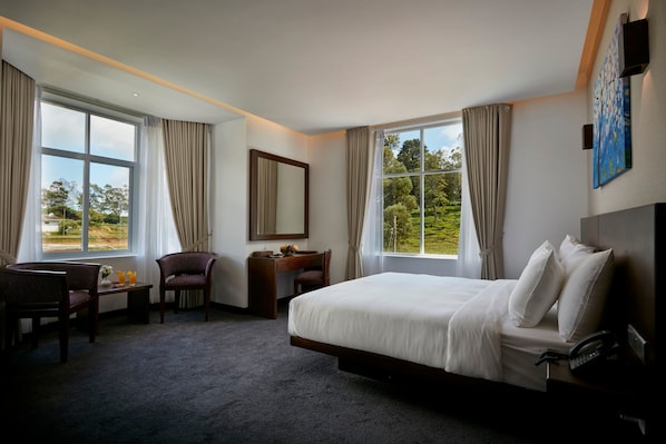 A hotel room with a large bed, a desk with a chair, a small table with a chair, and a large window with curtains.