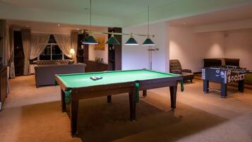 A billiards table, a foosball table, and a sofa in a room with a carpeted floor and a window with curtains.