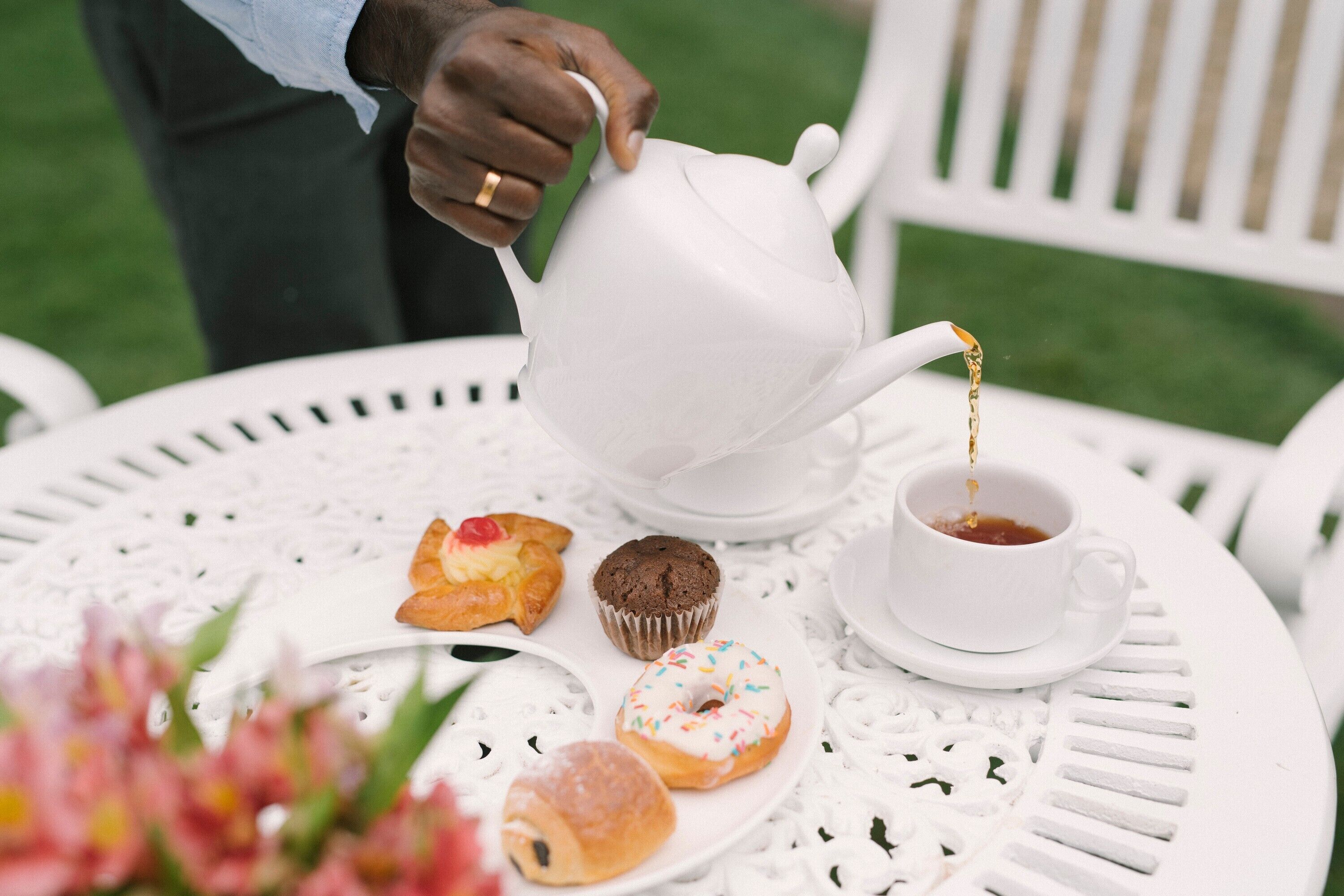 A white teapot pouring tea into a cup on a white table with pastries.