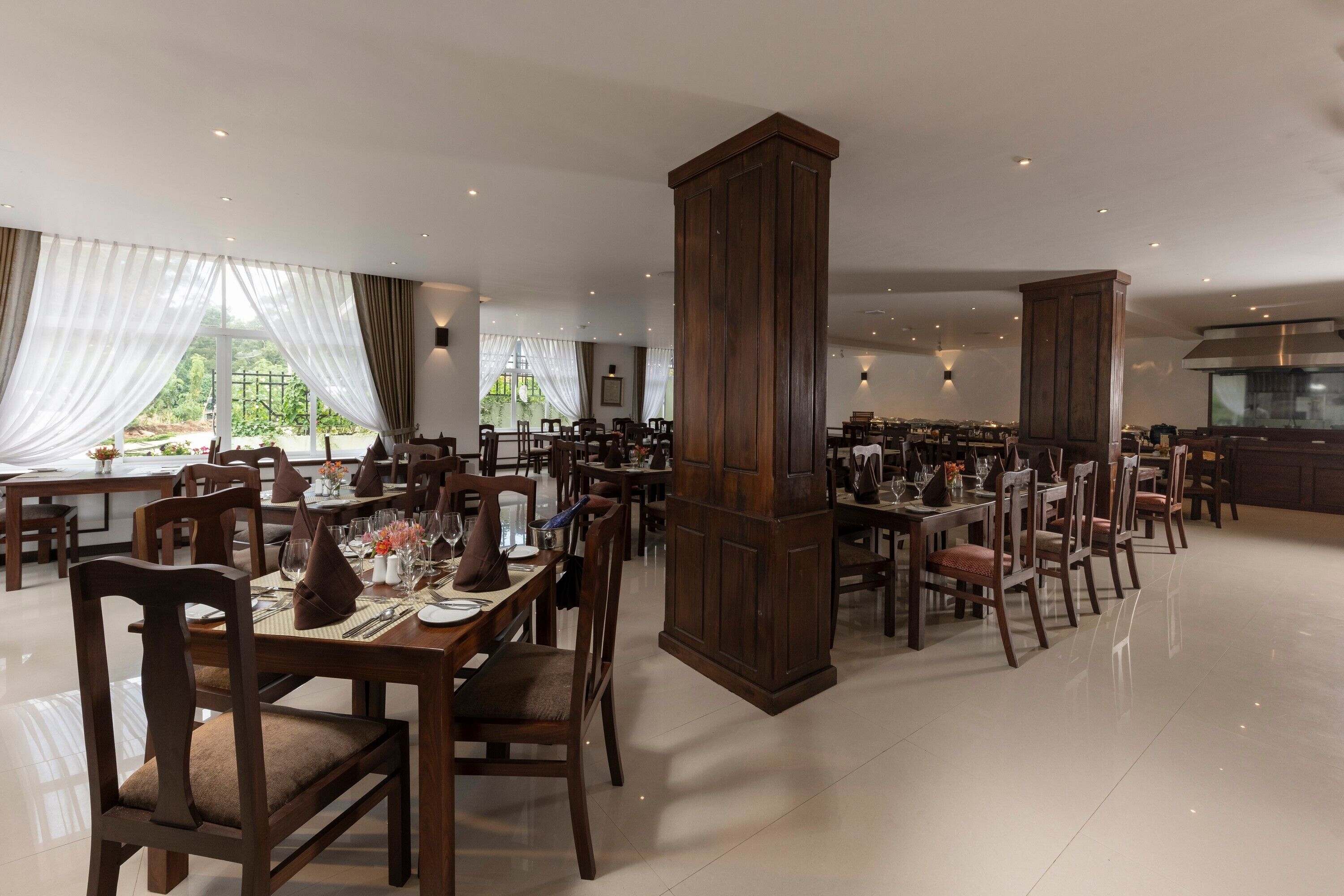 A spacious dining area with multiple tables and chairs, large wooden pillars, and a view of greenery outside.