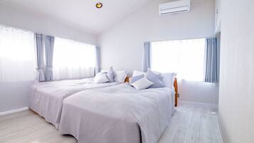 A modern bedroom with two beds, large windows with curtains, a ceiling light, and an air conditioning unit.