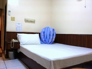 A single bed with a white mattress and a blue pillow, a bedside telephone, and a small trash bin.