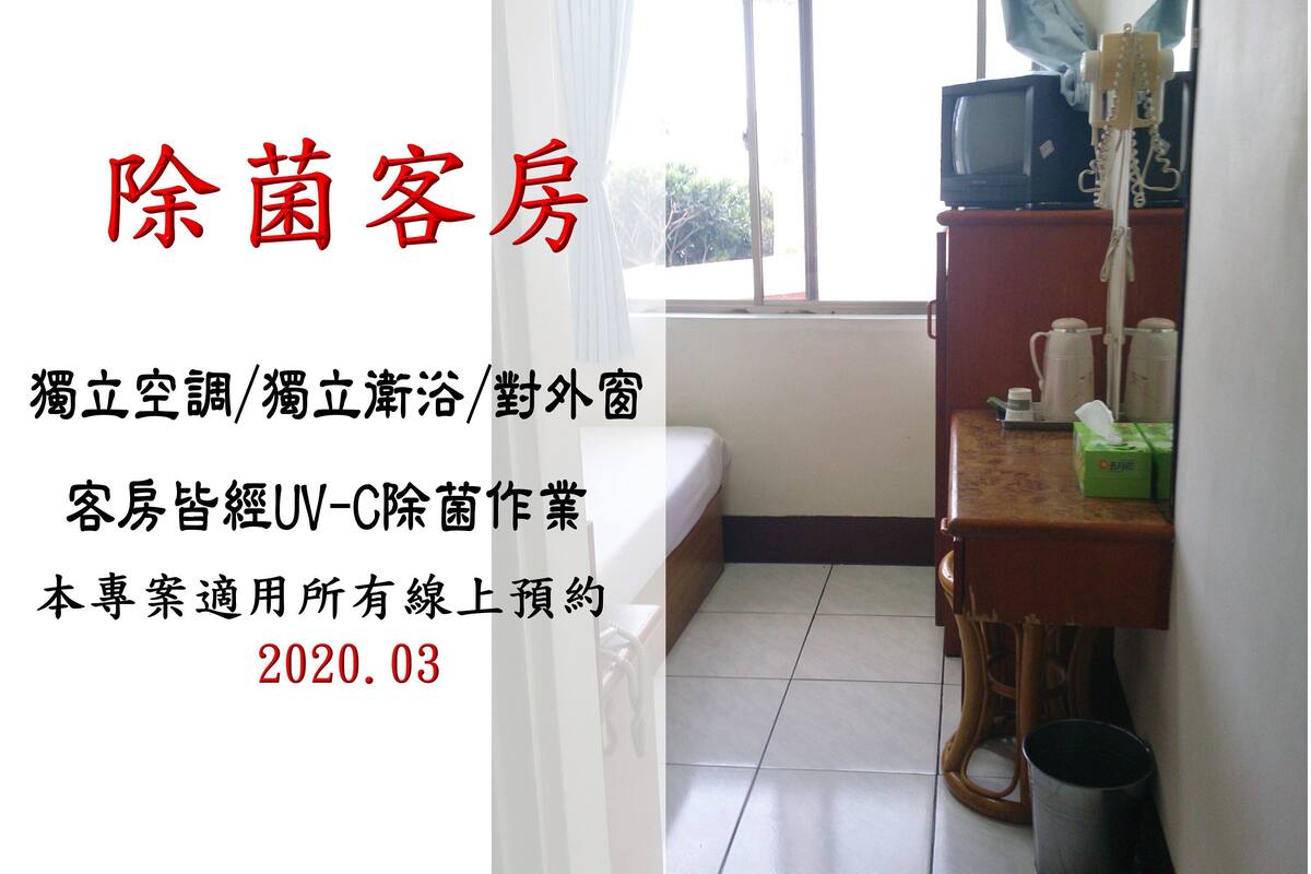 A hotel room with a bed, a small table with a television and some items on it, and a window with curtains.