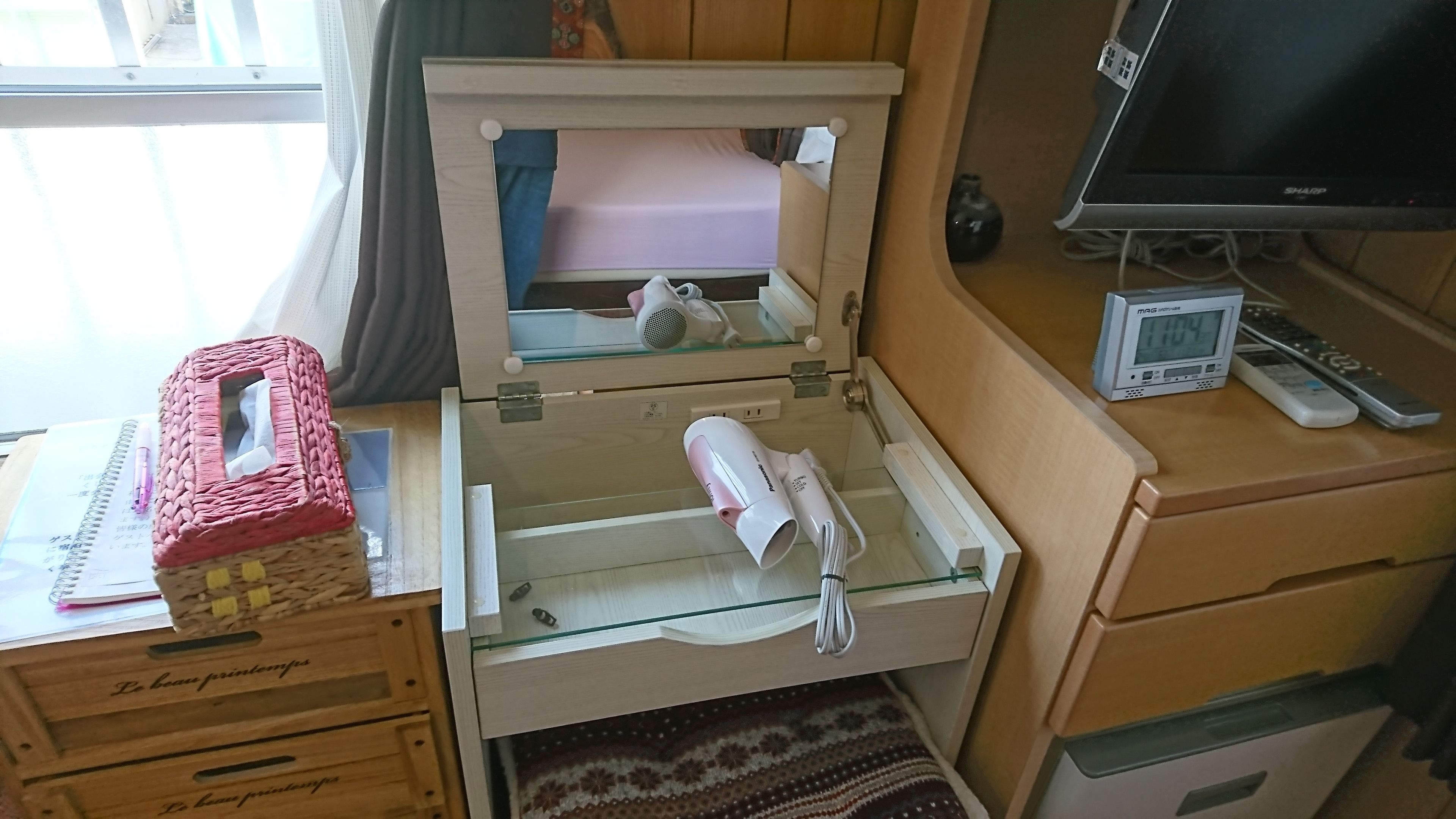 A compact bedroom with a bed, a small desk with a laptop, a bedside table with a lamp, and a window with curtains.