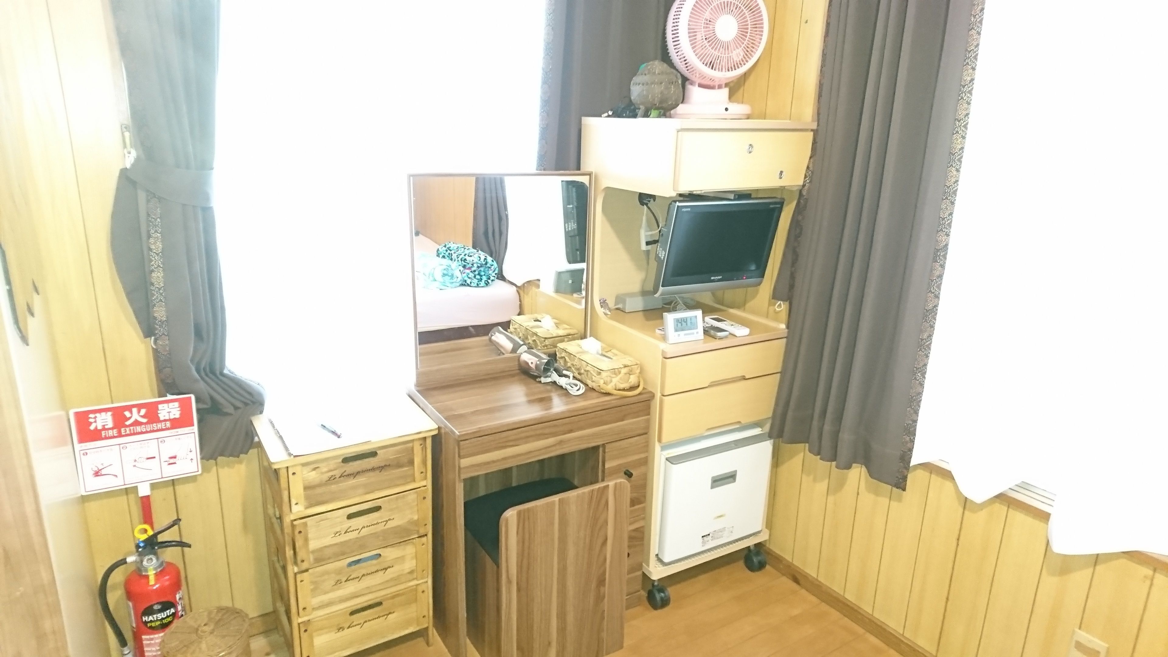 A compact room with a wooden desk, a computer, a fan, and a fire extinguisher.